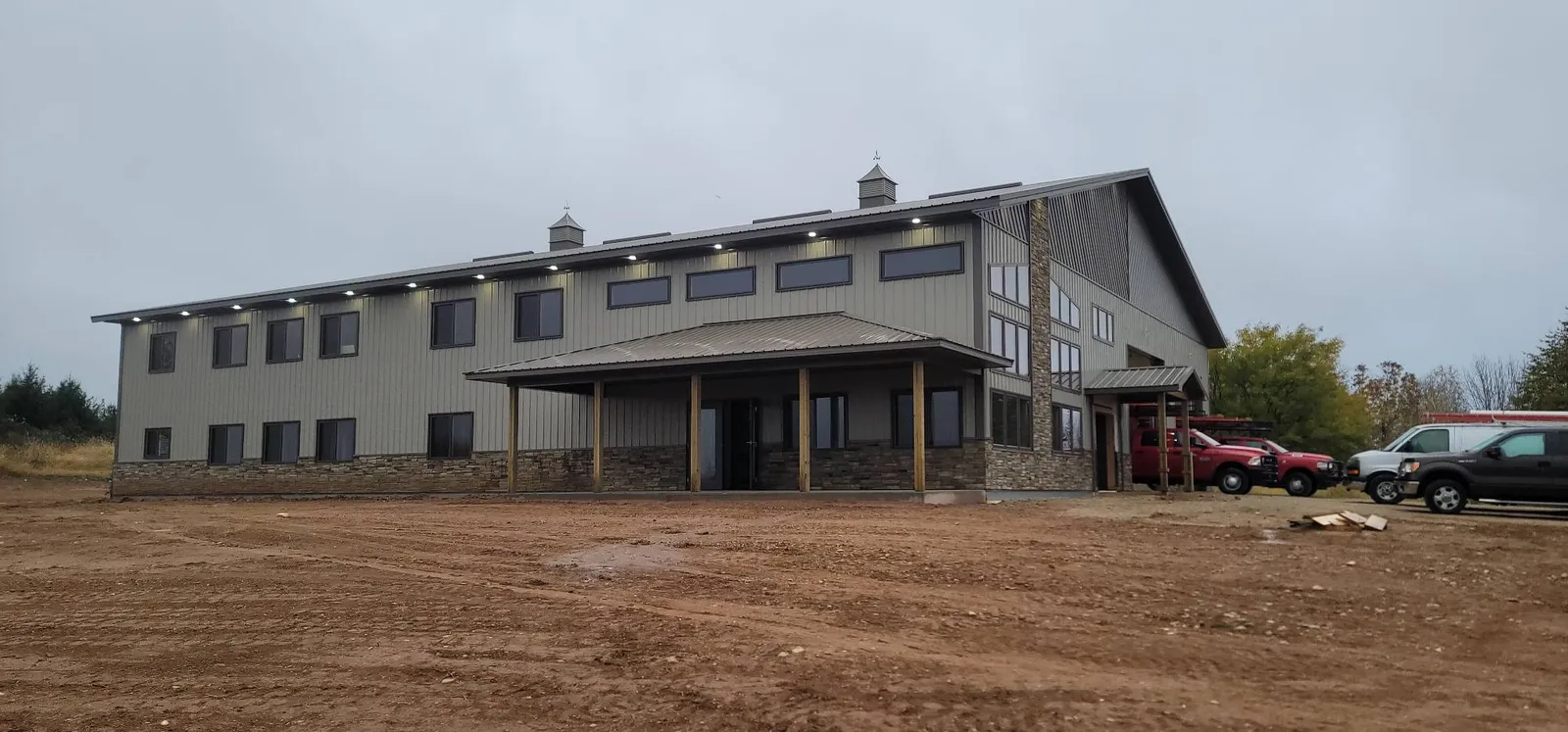 A two-storey post-frame barndominium with grey steel siding, stone wainscot and a covered porch, built by M.P.B. Builders